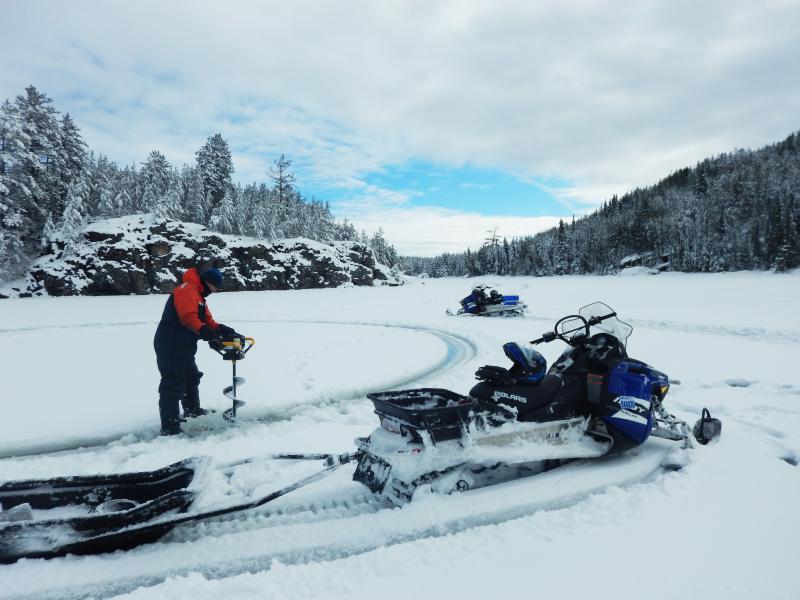 A snowy winter photo on an IISD-ELA lake, showing a snowmobile pulling a sled and a researcher drilling an ice corer into the frozen lake.